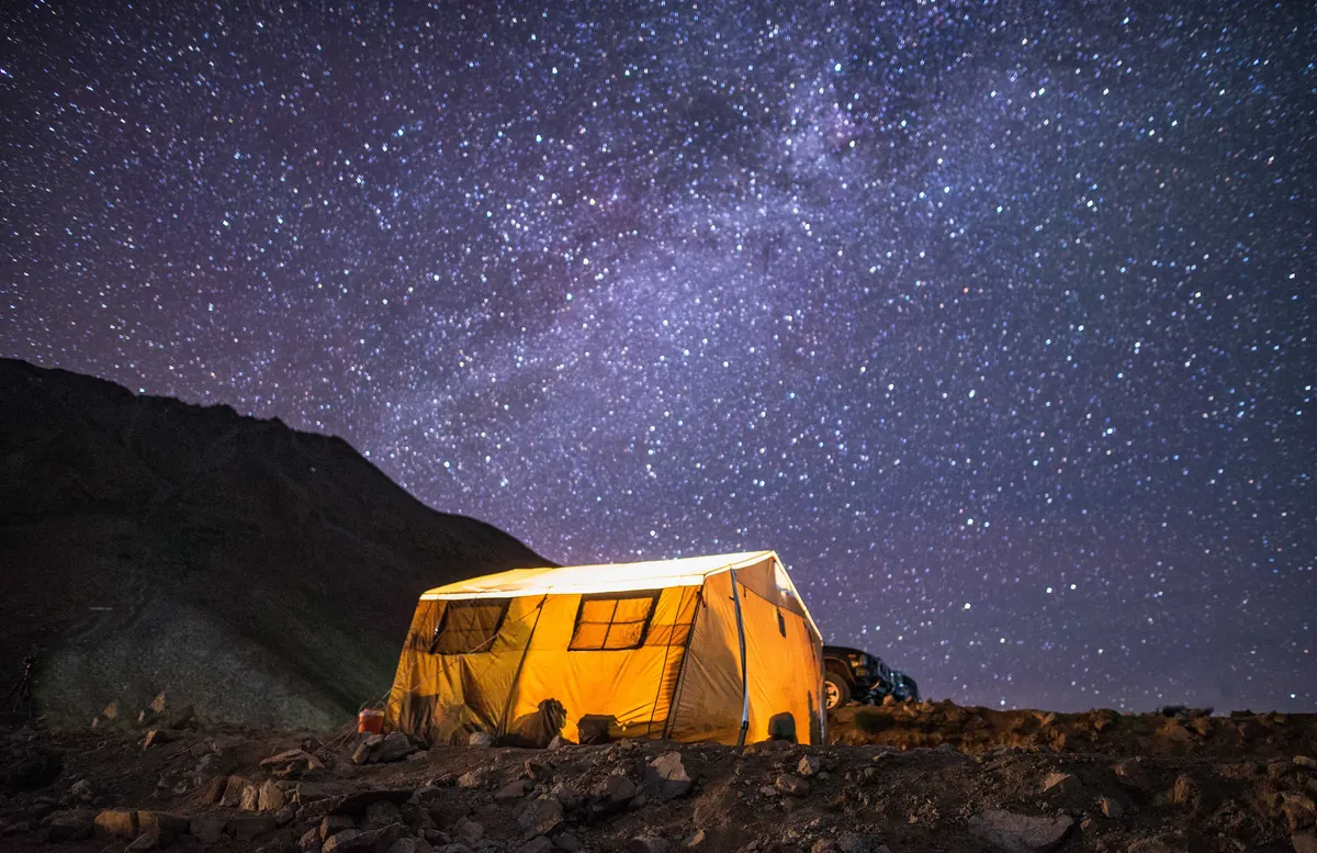 Tienda de campaña iluminada desde dentro en una llanura de alta montaña, bajo un cielo completamente estrellado